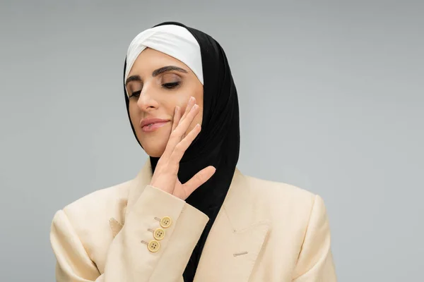 Expressive, stylish muslim businesswoman with closed eyes and makeup holding hand near chin on grey — Stock Photo