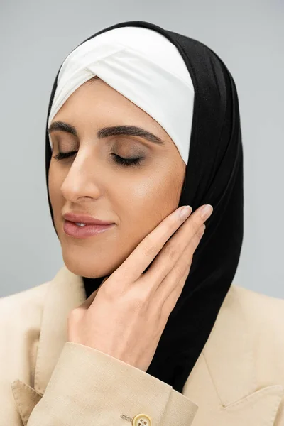 Porträt einer entzückten und eleganten muslimischen Geschäftsfrau mit geschlossenen Augen, die Gesicht auf Grau berührt — Stockfoto