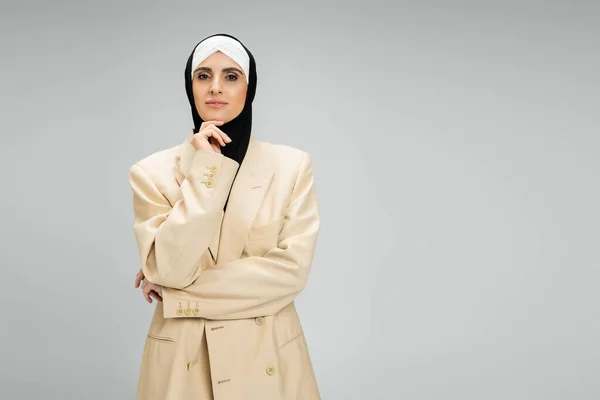 Ambitionierte muslimische Geschäftsfrau in Blazer und Hijab, die Hand am Kinn, in die Kamera blickend auf grau — Stockfoto