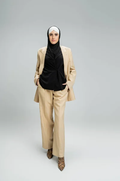 Сучасна мусульманська бізнес-леді в бежевому костюмі і хіджабі позує руками в кишенях на сірому — стокове фото