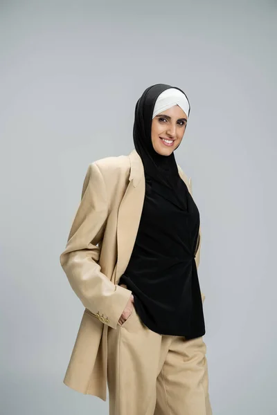 Alegre y de moda musulmana mujer de negocios en hijab y traje elegante beige mirando a la cámara en gris - foto de stock