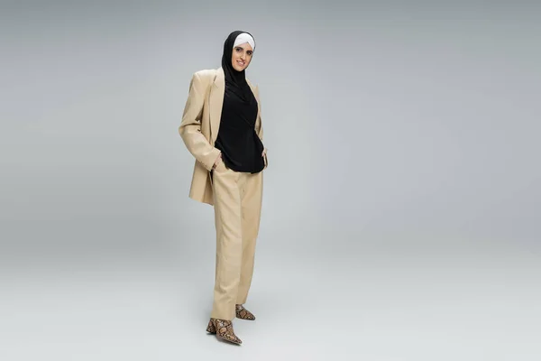 Fröhliche muslimische Geschäftsfrau in beigem Trendanzug und Hijab posiert mit den Händen in den Taschen auf grau — Stockfoto