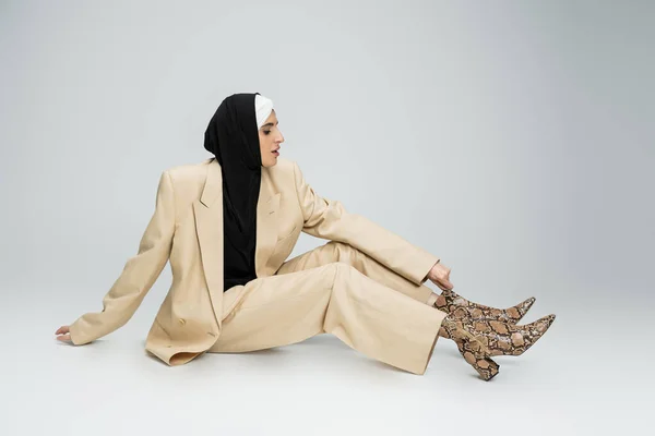 Trendy muslim businesswoman in hijab and beige suit adjusting snakeskin print boot on grey — Stock Photo