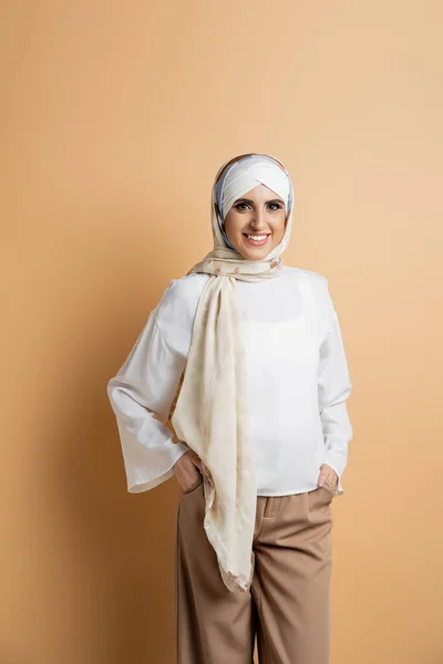 Mujer musulmana de moda en bufanda de seda y blusa blanca posando con las manos en bolsillos de pantalones en beige - foto de stock