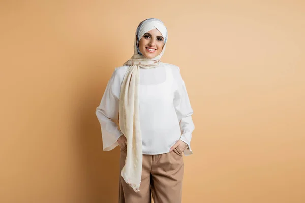 Mulher muçulmana alegre em blusa branca e lenço de cabeça de seda de pé com as mãos em bolsos em bege — Fotografia de Stock