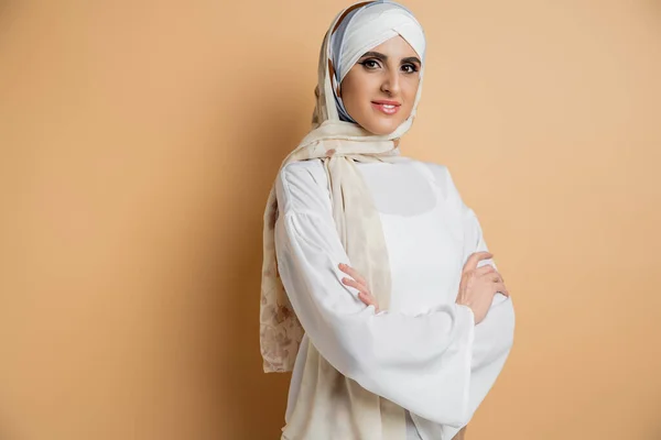 Positive muslim woman in silk headscarf and white blouse standing with folded arms on beige — Stock Photo