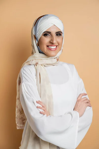 Modebewusste Muslimin in Frau mit strahlendem Lächeln, in stylischem Outfit, mit verschränkten Armen auf beige — Stockfoto