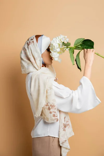 Muslim woman in elegant headscarf and blouse obscuring face with white hydrangea flower on beige — Stock Photo