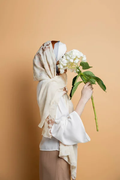 Elegante muslimische Frau in seidenem Schal und weißer Bluse, die das Gesicht mit Hortensienblume auf beige verdeckt — Stockfoto