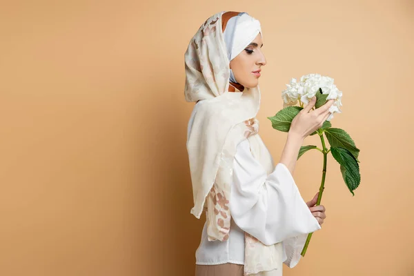 Gracieuse femme musulmane en foulard de soie et chemisier tenant fleur blanche sur beige, vue de côté — Photo de stock
