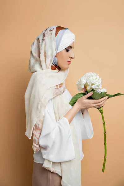 Side view of trendy muslim woman in headscarf and white blouse holding hydrangea flower on beige — Stock Photo