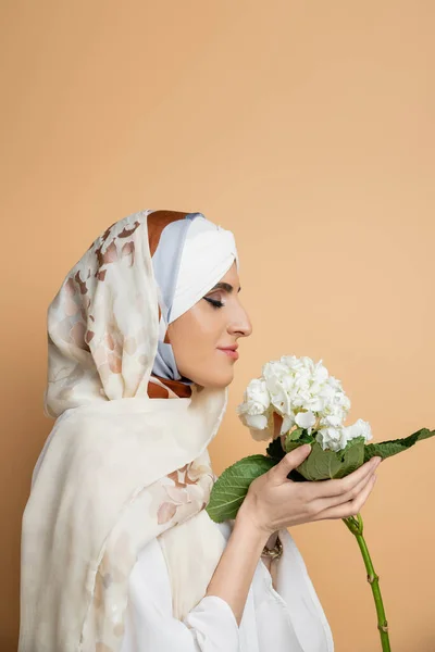 Delighted muslim woman in elegant headscarf holding white hydrangea flower on beige, side view — Stock Photo