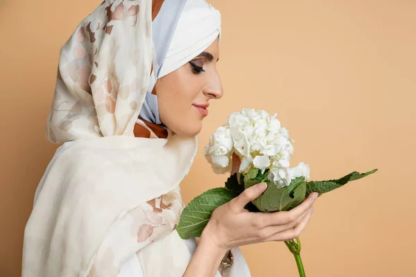 Smiling muslim woman in stylish headscarf holding white hydrangea flower on beige, closed eyes — Stock Photo