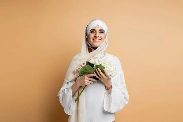 Восторженная мусульманка в блузке и платке держит цветок гортензии, смотрит в камеру на бежевый — стоковое фото