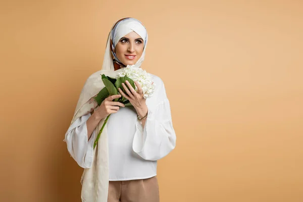 Hermosa mujer musulmana en pañuelo de seda y blusa de pie con flor de hortensia blanca en beige - foto de stock