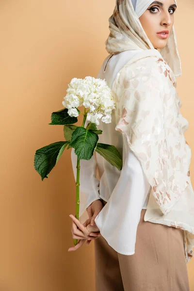 Charmante muslimische Frau mit seidenem Kopftuch, mit weißen Hortensienblüten, schaut in die Kamera auf beige — Stockfoto