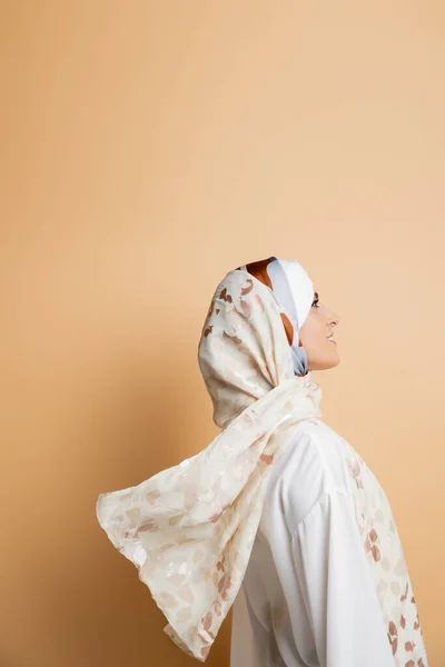 Vista lateral de mujer musulmana sonriente en elegante pañuelo de seda y blusa blanca en beige - foto de stock