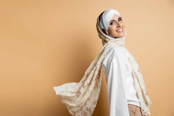 Радісна мусульманка в елегантних шовкових головних уборах і білій блузці дивиться на камеру на бежевому — стокове фото
