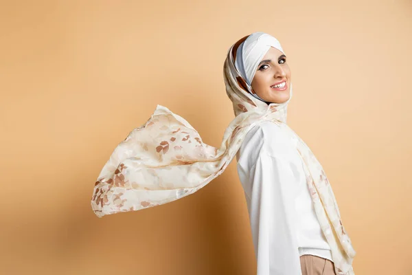 Carefree and stylish muslim woman in waving silk headscarf looking at camera on beige — Stock Photo