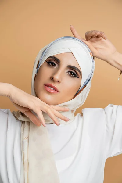 Encantadora mujer musulmana con maquillaje posando en pañuelo de seda y blusa blanca en beige, retrato - foto de stock