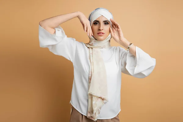 Елегантна мусульманка в білій блузці і шовкові головикарф позує руками біля обличчя на бежевому — стокове фото