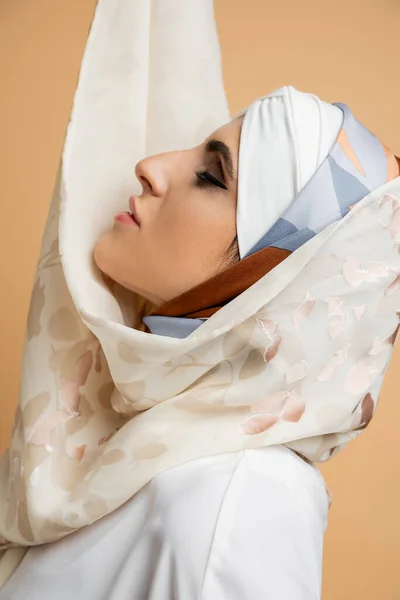 Профіль чарівної мусульманської жінки з макіяжем позує в стильному шовковому шарфі на бежевому — стокове фото