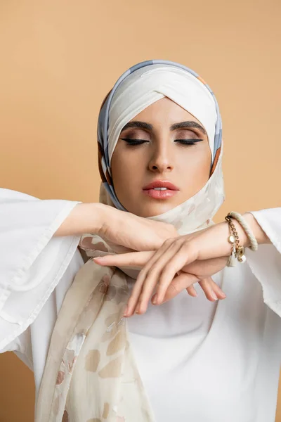 Mujer musulmana de moda con maquillaje y ojos cerrados posando en bufanda de seda y blusa blanca en beige - foto de stock
