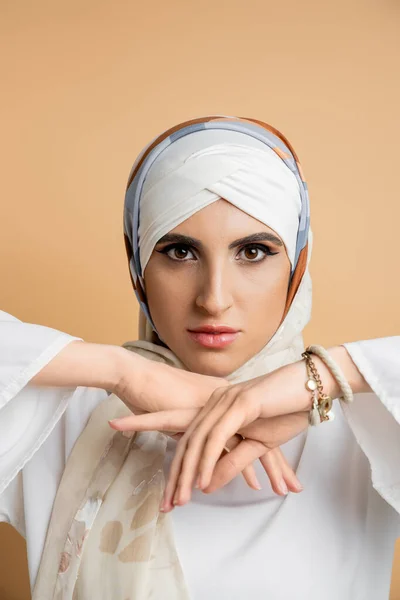 Encantadora mulher do Oriente Médio em elegante cachecol de seda olhando para a câmera em bege, beleza muçulmana — Fotografia de Stock