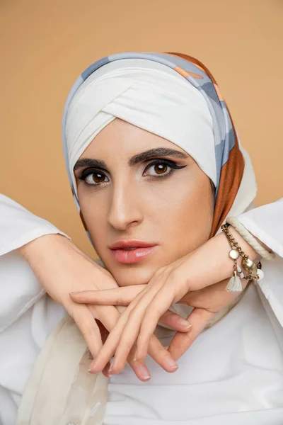 Portrait of glamour muslim woman with makeup posing in silk scarf with hands near chin on beige — Stock Photo