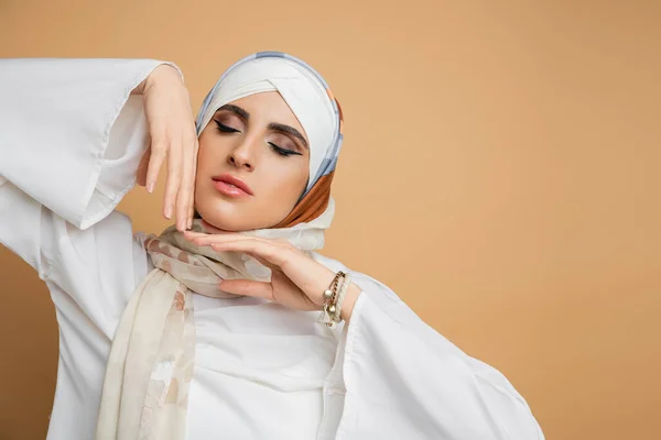 Portrait de femme musulmane sensuelle en écharpe de soie posant les yeux fermés et les mains près du visage sur beige — Photo de stock