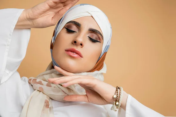 Bela mulher muçulmana com as mãos perto do rosto e olhos fechados em cachecol de seda no bege, retrato — Fotografia de Stock