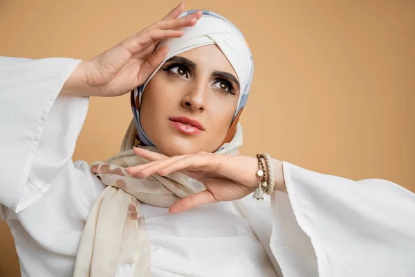 Mulher muçulmana elegante em cachecol de seda e blusa branca posando com as mãos perto do rosto no bege — Fotografia de Stock