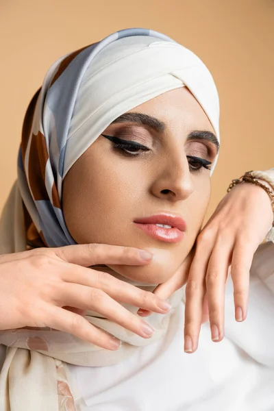 Portrait of modern muslim woman with makeup and hands near face posing in silk headscarf on beige — Stock Photo