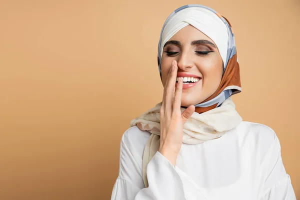 Joyful muslim woman with makeup wearing elegant attire and laughing with closed eyes on beige — Stock Photo