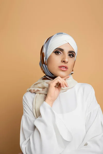 Sensual muslim woman in stylish outfit posing with hand near chin and looking at camera on beige — Stock Photo