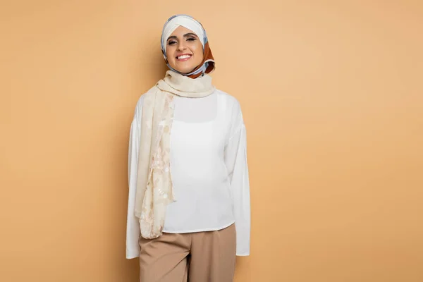 Alegre mujer musulmana en blusa blanca y bufanda de seda de pie y sonriendo a la cámara en beige - foto de stock