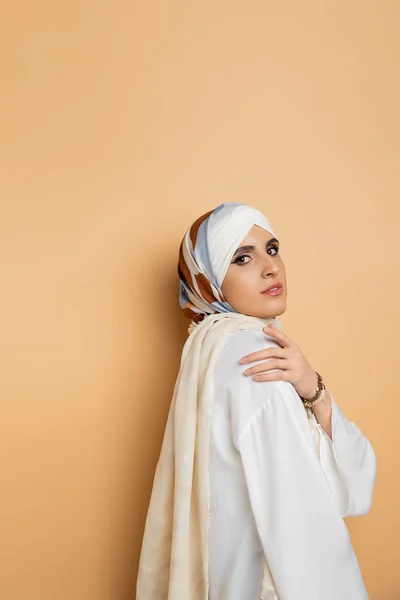 Charmante muslimische Frau in weißer Bluse und Seidentuch blickt in die Kamera auf beige, Schönheit und Stil — Stockfoto