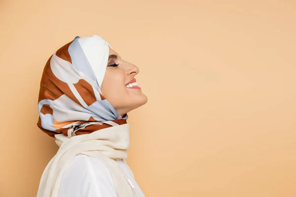 Side view of fashionable muslim woman in silk headscarf smiling with closed eyes on beige — Stock Photo