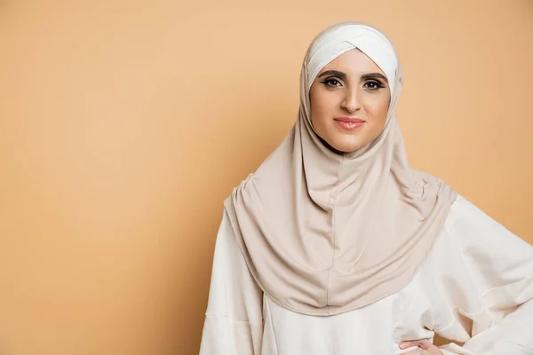 Positive muslim woman in hijab and sweatshirt posing with hand on waist, looking at camera on beige — Stock Photo