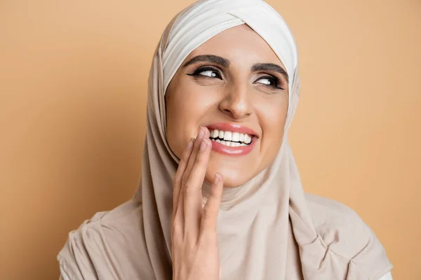 Atractiva, mujer musulmana llena de alegría con maquillaje usando hijab, riendo y mirando hacia otro lado en beige - foto de stock