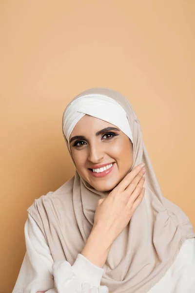 Compiaciuta donna musulmana con il trucco indossando hijab e sorridendo con mano vicino al viso sul beige, ritratto — Foto stock
