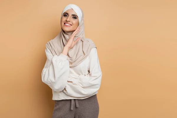 Glückliche muslimische Frau im Hijab und trendiger Freizeitkleidung, die mit händennahem Gesicht auf beige in die Kamera blickt — Stockfoto