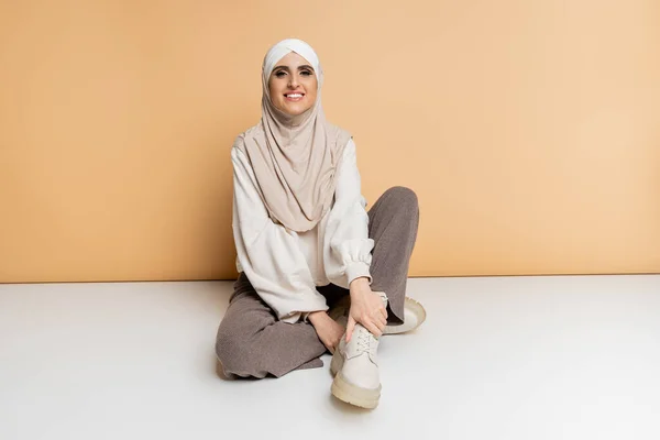 Happy muslim woman in hijab and modern casual attire sitting on beige, full length, personal style — Stock Photo
