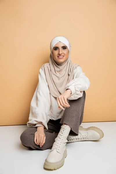 Muslim woman in hijab, white sweatshirt, grey pants and leather boots sitting and smiling on beige — Stock Photo