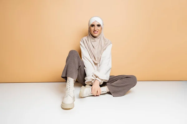 Mulher muçulmana alegre em hijab, calças e botas de couro sentado e olhando para a câmera no bege — Fotografia de Stock