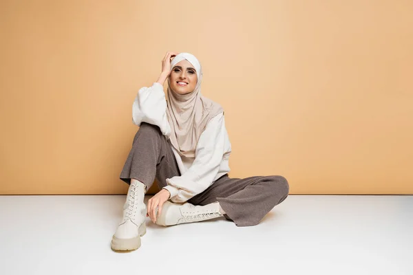 Mulher muçulmana na moda em hijab, camisola, calças e botas sentado e sorrindo para a câmera no bege — Fotografia de Stock