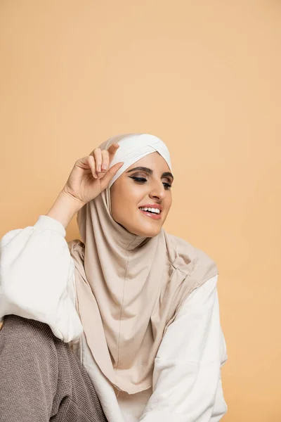 Femme musulmane charmante et heureuse avec maquillage, en hijab et sweat blanc assis sur beige — Photo de stock