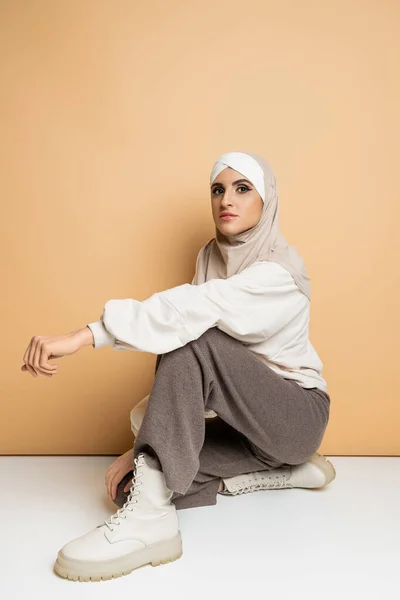 Mulher muçulmana confiante em hijab, calças cinzentas e botas de couro sentado e olhando para a câmera no bege — Fotografia de Stock