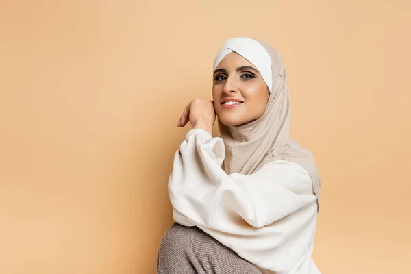 Modern muslim woman with makeup wearing hijab and white sweatshirt while smiling at camera on beige — Stock Photo
