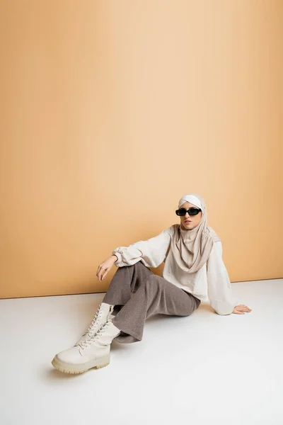 Модная мусульманка в хиджабе, солнцезащитных очках и современной повседневной одежде, сидящая на бежевом, индивидуальном стиле — стоковое фото